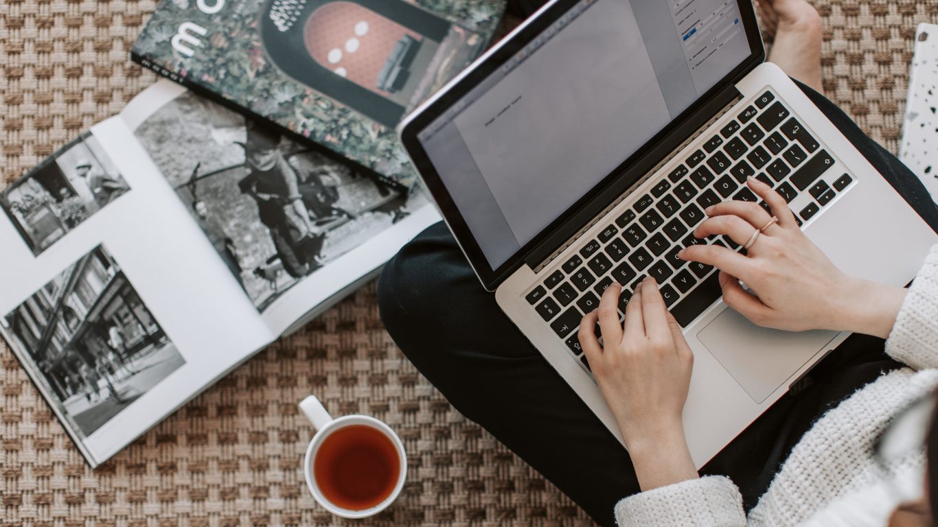 An open laptop, an open magazine and a cup of tea laid out on a rug.
